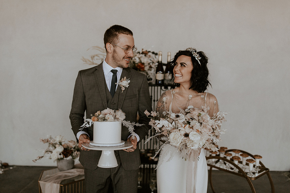  bride in a formfitting satin gown with a sheer cape and the groom in a café brown suit and chocolate long tie and the bridesmiads in white gowns