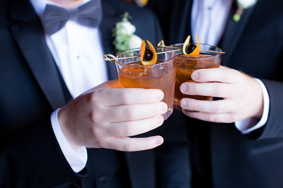 grooms in black tuxedos with white shirts and black bow ties holding cocktails