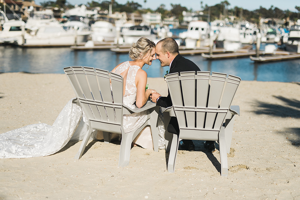 bride in a white flowing gown with deep v neck line and thin straps, the groom is in a black tuxedo sitting in chairs by the ocean