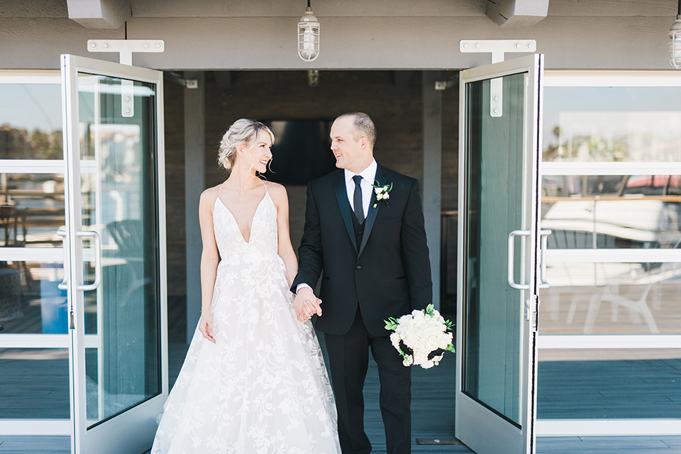 bride in a white flowing gown with deep v neck line and thin straps, the groom is in a black tuxedo standing in the doorway of venue