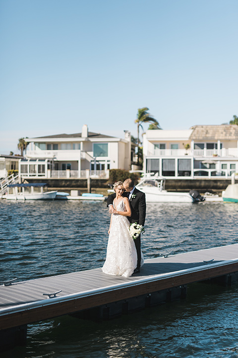 bride in a white gown with a deep v neckline and thin straps and the groom in a black tuxedo on the dock