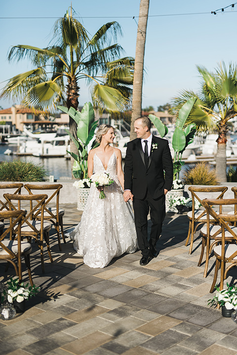 bride in a white flowing gown with thin straps and hair in a lose bun, the groom in a black tuxedo and black long tie- walking down the ceremony aisle together