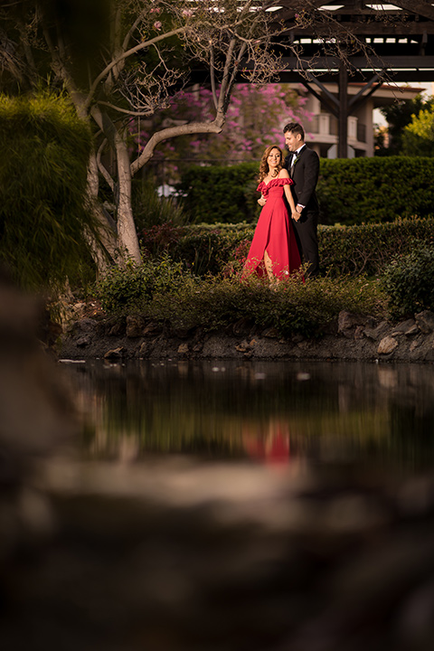  girl in a long red dress with an off the shoulder detail and roses and man in a black tuxedo with a shawl lapel tuxedo and a black bow tie dancing in the park