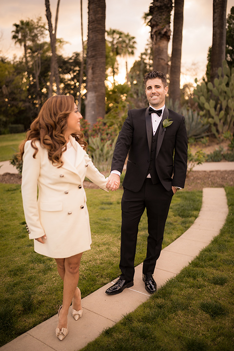  girl in a long cream trench coat and man in a black tuxedo with a shawl lapel tuxedo and a black bow tie dancing in the park