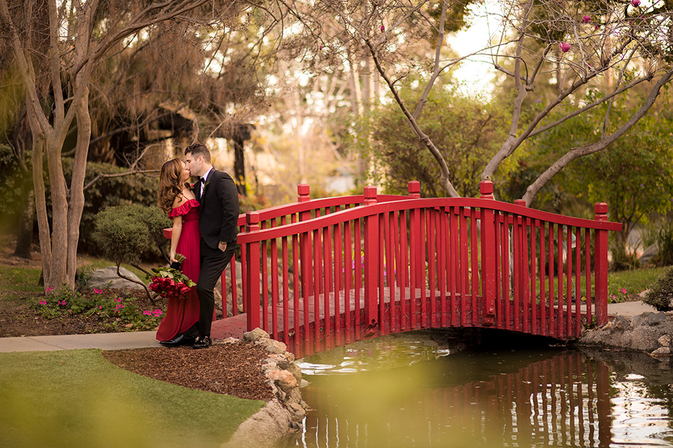  girl in a long red dress with an off the shoulder detail and roses and man in a black tuxedo with a shawl lapel tuxedo and a black bow tie by the bridge 