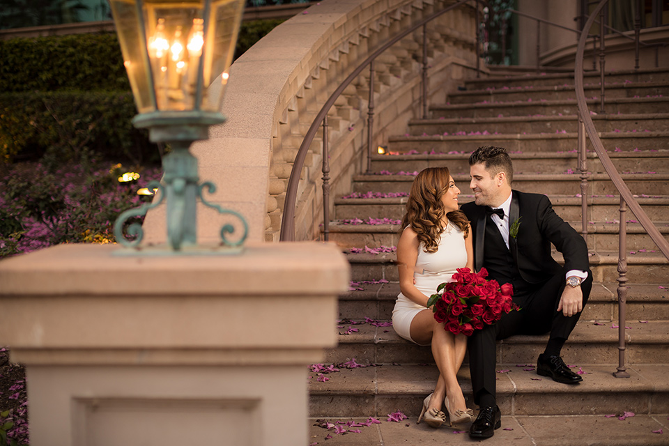  girl in a long cream colored trench coat and man in a black tuxedo with a shawl lapel tuxedo and a black bow tie sitting on the steps 