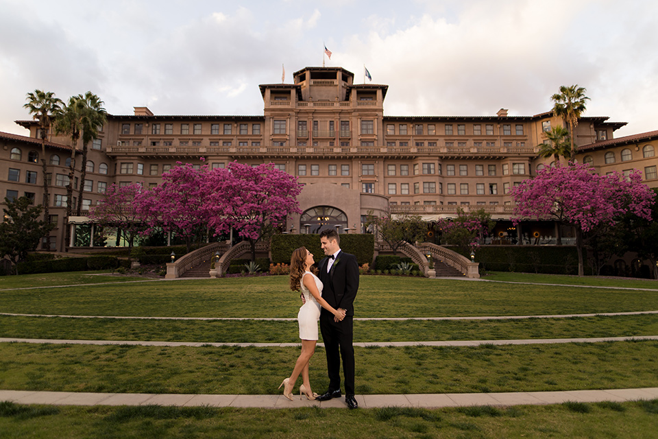  girl in a long cream colored trench coat and man in a black tuxedo with a shawl lapel tuxedo and a black bow standing outside the venue 