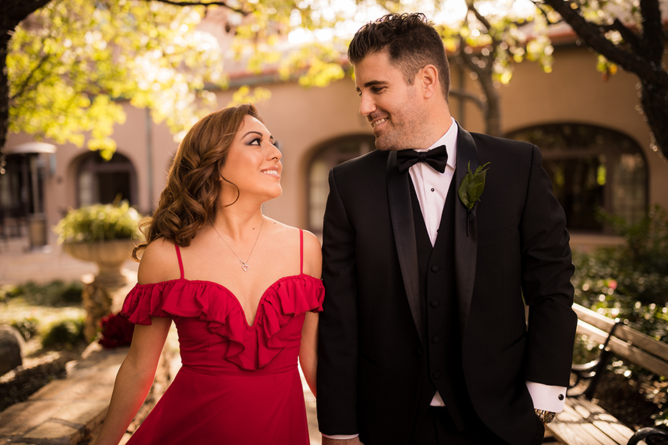  girl in a long red dress with an off the shoulder detail and roses and man in a black tuxedo with a shawl lapel tuxedo and a black bow tie walking in the park 