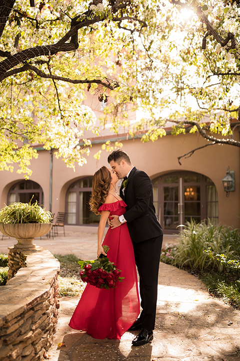  girl in a long red dress with an off the shoulder detail and roses and man in a black tuxedo with a shawl lapel tuxedo and a black bow tie kissing in the park