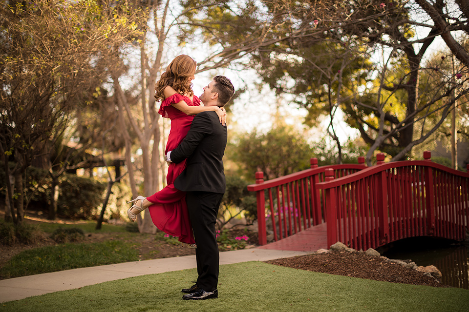  girl in a long red dress with an off the shoulder detail and roses and man in a black tuxedo with a shawl lapel tuxedo and a black bow tie walking in the park 
