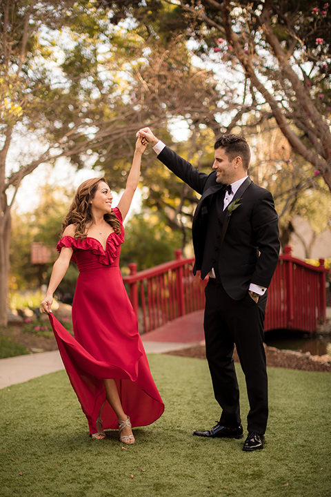 girl in a long red dress with an off the shoulder detail and roses and man in a black tuxedo with a shawl lapel tuxedo and a black bow tie twirling