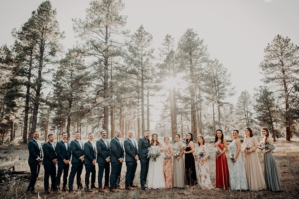  groom and groomsmen in dark blue suits with bow ties, bridesmaids in soft pastel colors in different patterns and designs, the bride in a flowing white gown with an off the shoulder detail 