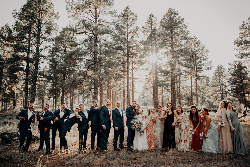  groom and groomsmen in dark blue suits with bow ties, bridesmaids in soft pastel colors in different patterns and designs, the bride in a flowing white gown with an off the shoulder detail 