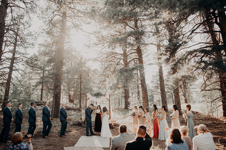  groom and groomsmen in dark blue suits with bow ties, bridesmaids in soft pastel colors in different patterns and designs, the bride in a flowing white gown with an off the shoulder detail 