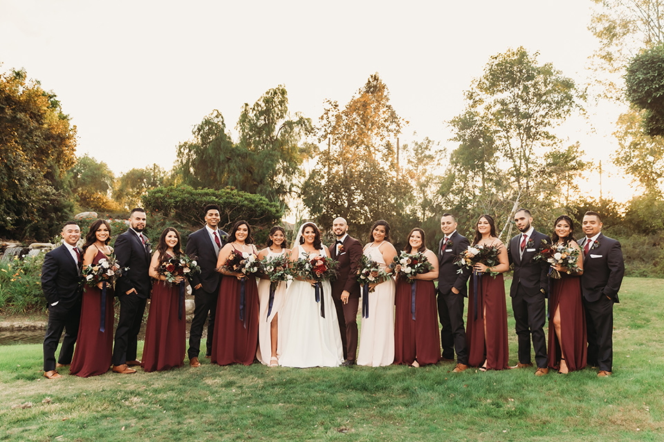  bride in a white ballgown with a plunging neckline and long veil and the groom in a burgundy tuxedo with a black shawl lapel, the bridesmaids in burgundy gowns and the groomsmen in black tuxedos