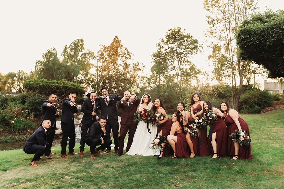  bride in a white ballgown with a plunging neckline and long veil and the groom in a burgundy tuxedo with a black shawl lapel, the bridesmaids in burgundy gowns and the groomsmen in black tuxedos