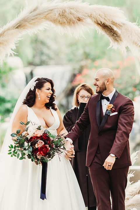  bride in a white ballgown with a plunging neckline and long veil and the groom in a burgundy tuxedo with a black shawl lapel 