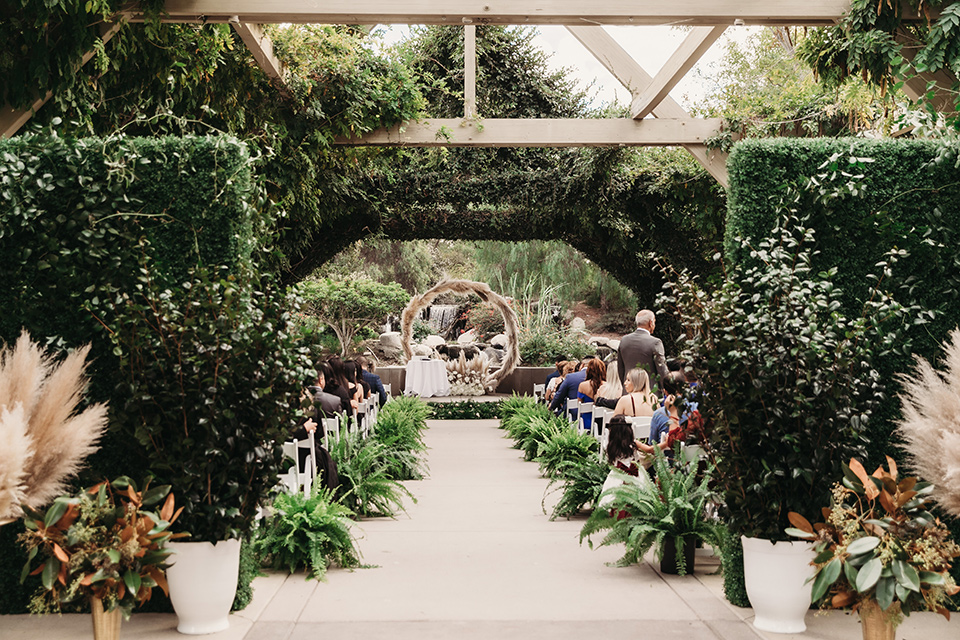  bride in a white ballgown with a plunging neckline and long veil and the groom in a burgundy tuxedo with a black shawl lapel, the bridesmaids in burgundy gowns and the groomsmen in black tuxedos with pampas grass arch at the ceremony