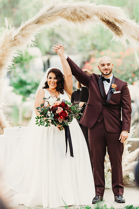  bride in a white ballgown with a plunging neckline and long veil and the groom in a burgundy tuxedo with a black shawl lapel