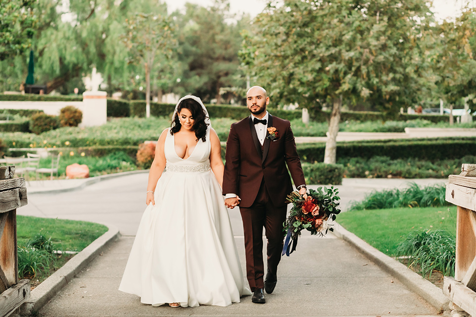  bride in a white ballgown with a plunging neckline and long veil and the groom in a burgundy tuxedo with a black shawl lapel, the bridesmaids in burgundy gowns and the groomsmen in black tuxedos