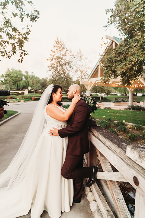  bride in a white ballgown with a plunging neckline and long veil and the groom in a burgundy tuxedo with a black shawl lapel