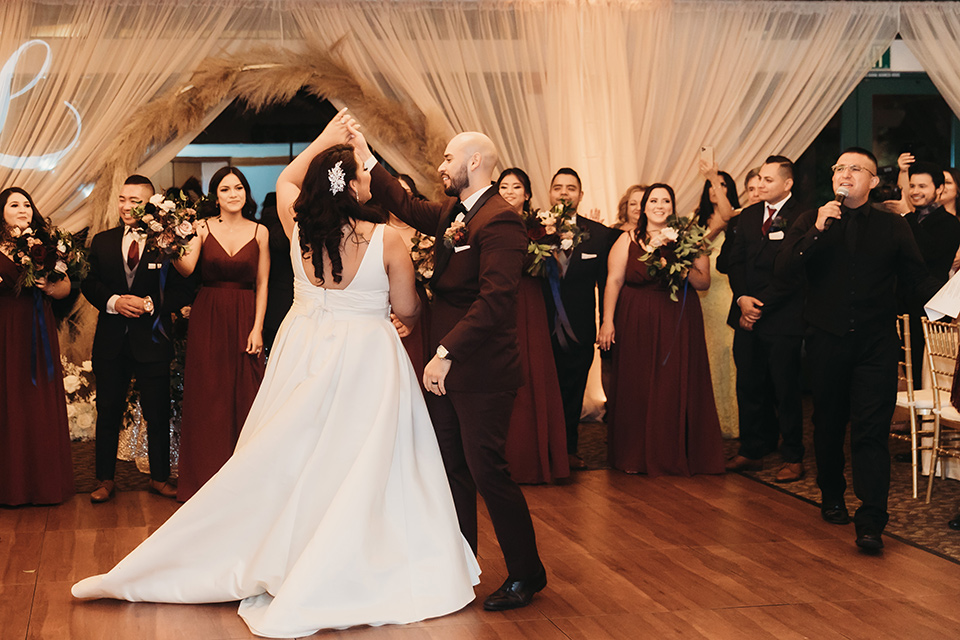  bride in a white ballgown with a plunging neckline and long veil and the groom in a burgundy tuxedo with a black shawl lapel, the bridesmaids in burgundy gowns and the groomsmen in black tuxedos