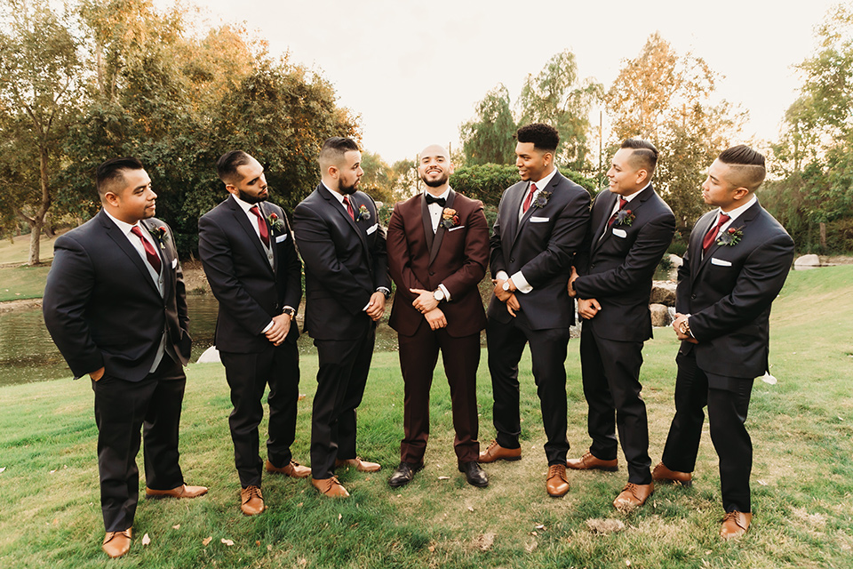  the groom in a burgundy tuxedo with a black shawl lapel the groomsmen in dark navy suits 