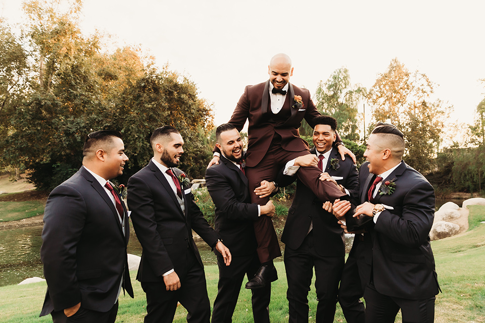  the groom in a burgundy tuxedo with a black shawl lapel the groomsmen in dark navy suits 