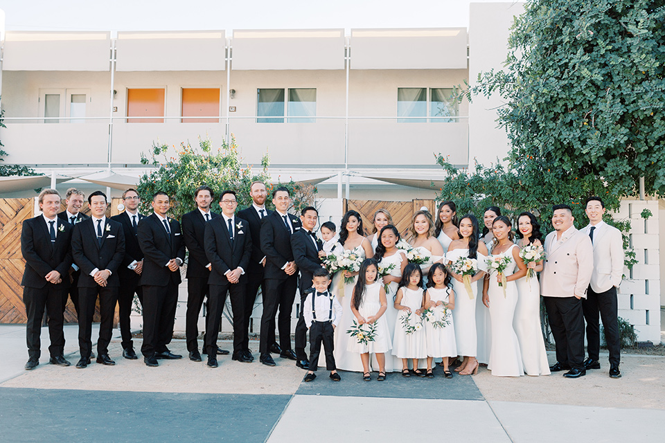  bride in a white modern gown with a low cut neckline and pockets and the groom in a black tuxedo and bow tie, the bridesmaids in neutral colored gowns and groomsmen in black tuxedos