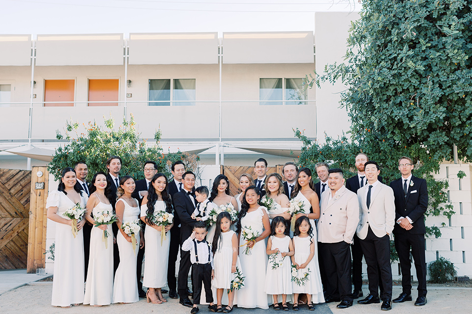  bride in a white modern gown with a low cut neckline and pockets and the groom in a black tuxedo and bow tie, the bridesmaids in neutral colored gowns and groomsmen in black tuxedos