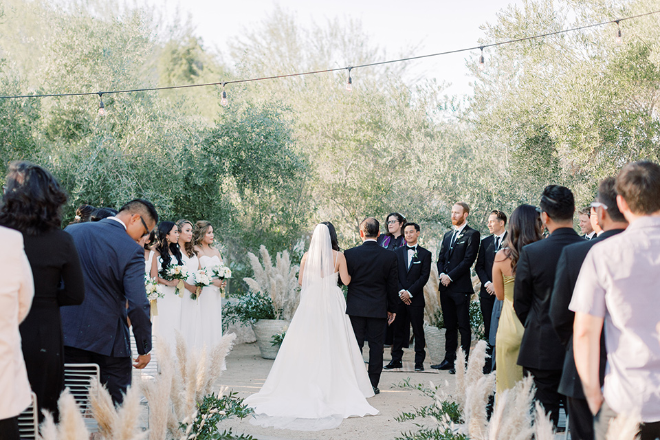  bride in a white modern gown with a low cut neckline and pockets and the groom in a black tuxedo and bow tie, the bridesmaids in neutral colored gowns and groomsmen in black tuxedos at the ceremony