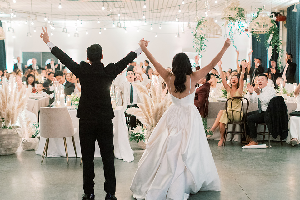  bride in a white modern gown with a low cut neckline and pockets and the groom in a black tuxedo and bow tie, the bridesmaids in neutral colored gowns and groomsmen in black tuxedos at the reception