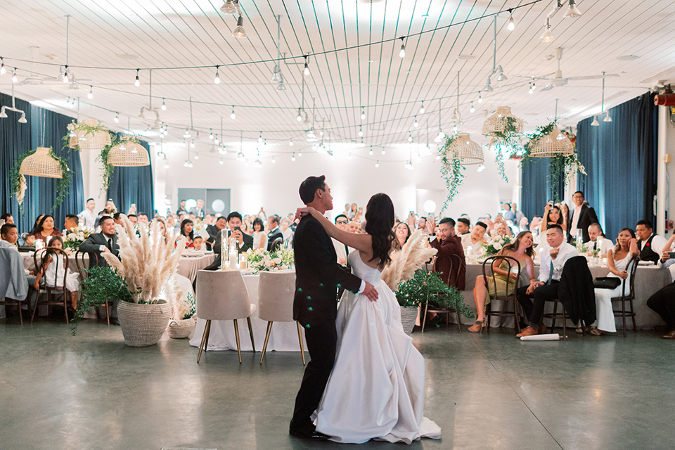  bride in a white modern gown with a low cut neckline and pockets and the groom in a black tuxedo and bow tie, the bridesmaids in neutral colored gowns and groomsmen in black tuxedos at the reception