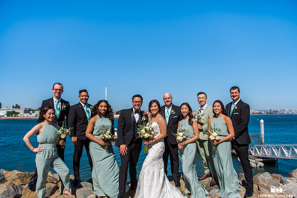  bride in a white lace form fitting gown with a strapless sweetheart neckline, the groom in a black tuxedo with a black bow tie, the groomsmen in black tuxedos with teal bow ties, the the bridesmaids in mint blue gowns and the bridesman in a mint suit to match the girls’ gown