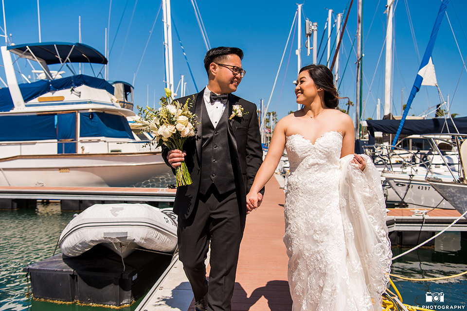  bride in a white lace form fitting gown with a strapless sweetheart neckline, the groom in a black tuxedo with a black bow tie, by the boats in the marina