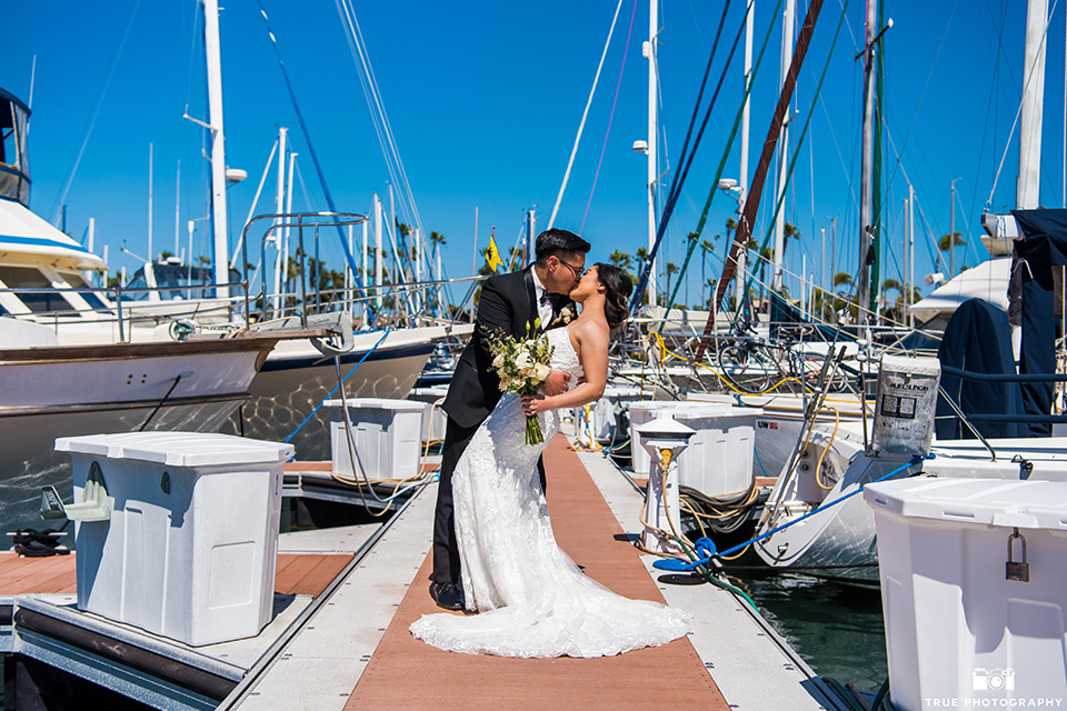  bride in a white lace form fitting gown with a strapless sweetheart neckline, the groom in a black tuxedo with a black bow tie, by the boats in the marina