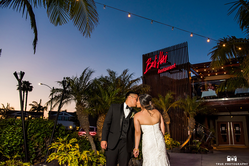  bride in a white lace form fitting gown with a strapless sweetheart neckline, the groom in a black tuxedo with a black bow tie, outside venue