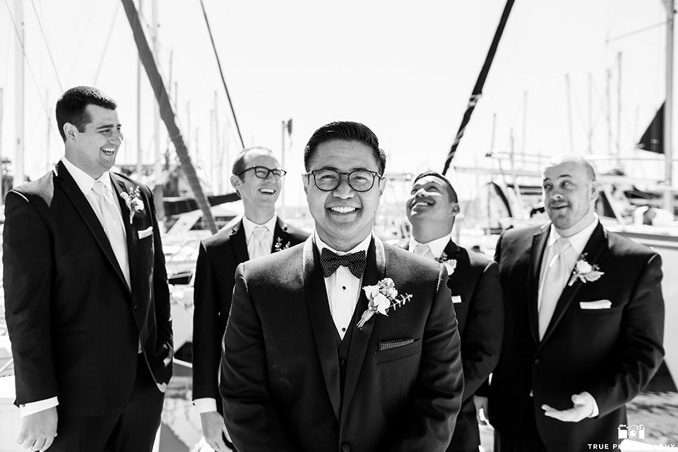  the groom in a black tuxedo with a black bow tie, the groomsmen in black tuxedos with teal bow ties