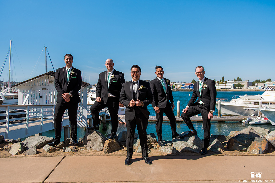  the groom in a black tuxedo with a black bow tie, the groomsmen in black tuxedos with teal bow ties