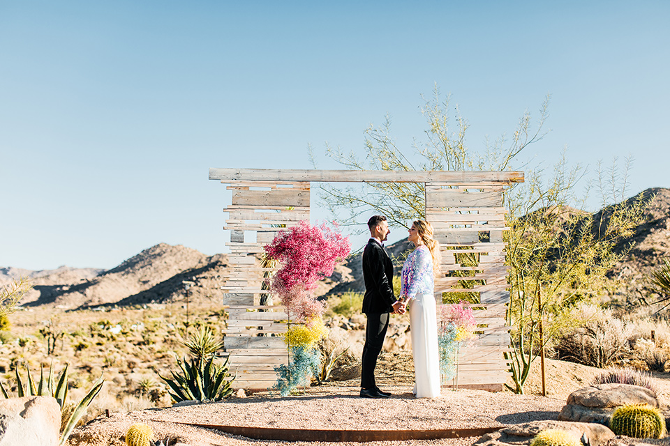  bride was in a white strapless jumpsuit with a colorful jacket and hair in a ponytail and the groom in a black paisley tuxedo with a peak lapel and black bow tie 