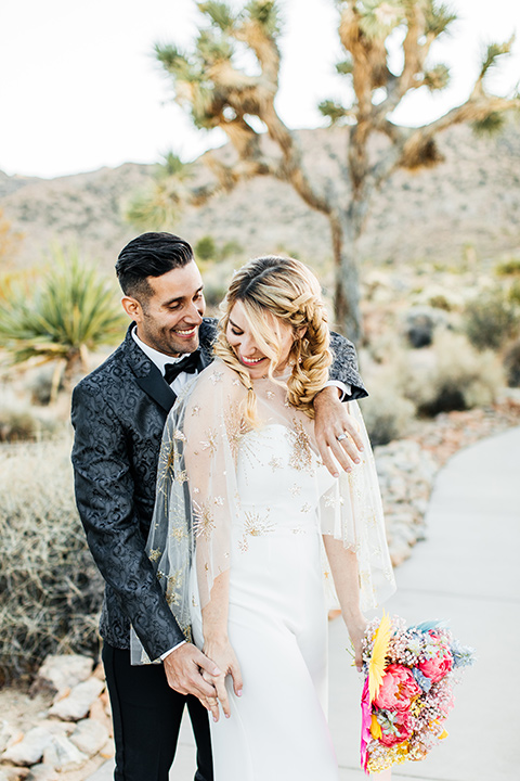  bride was in a white strapless jumpsuit with a colorful jacket and hair in a ponytail and the groom in a black paisley tuxedo with a peak lapel and black bow tie 