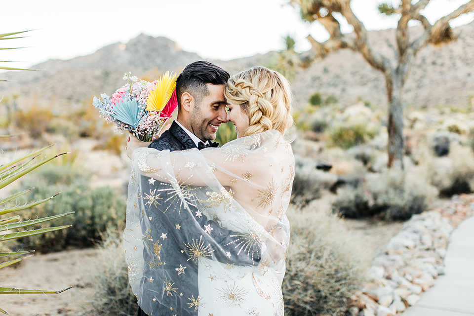  bride was in a white strapless jumpsuit with a colorful jacket and hair in a ponytail and the groom in a black paisley tuxedo with a peak lapel and black bow tie 