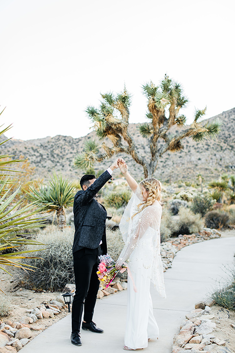  bride was in a white strapless jumpsuit with a colorful jacket and hair in a ponytail and the groom in a black paisley tuxedo with a peak lapel and black bow tie 