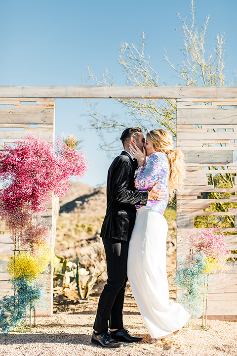  bride was in a white strapless jumpsuit with a colorful jacket and hair in a ponytail and the groom in a black paisley tuxedo with a peak lapel and black bow tie 