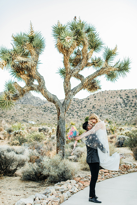  bride was in a white strapless jumpsuit with a colorful jacket and hair in a ponytail and the groom in a black paisley tuxedo with a peak lapel and black bow tie 