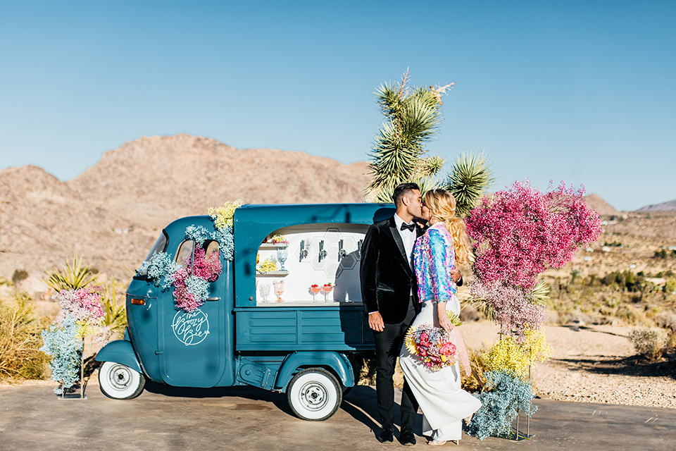  bride was in a white strapless jumpsuit with a colorful jacket and hair in a ponytail and the groom in a black paisley tuxedo with a peak lapel and black bow tie 