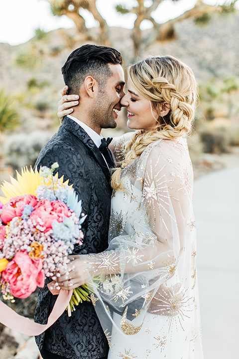  bride was in a white strapless jumpsuit with a colorful jacket and hair in a ponytail and the groom in a black paisley tuxedo with a peak lapel and black bow tie 