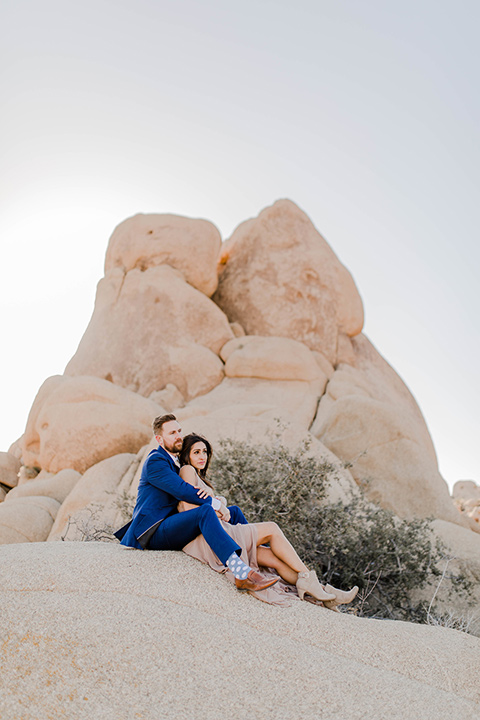 bride in a taupe maxi gown with straps and her hair down, the groom in a cobalt blue suit with a neutral long tie, sitting on dune
