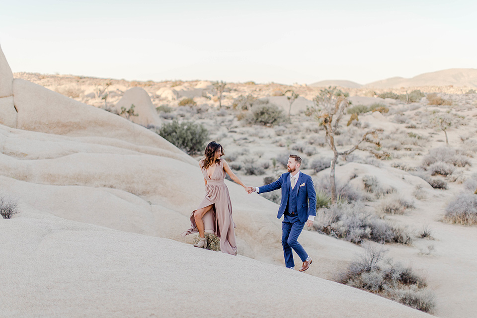  bride in a taupe gown with ankle boots and straps, the groom in a cobalt blue suit with a neutral colored tie walking up a sand dune