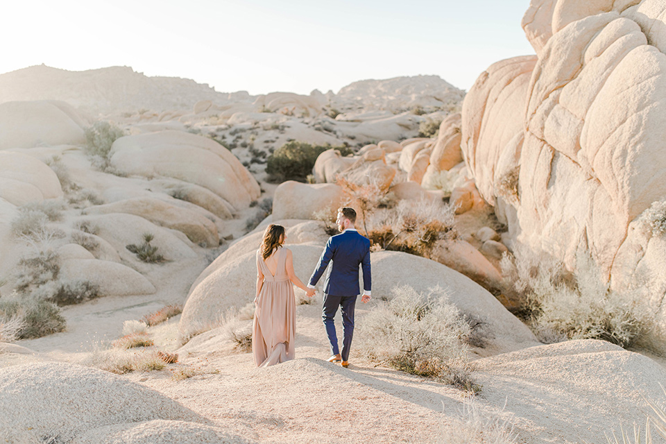  bride in a taupe gown with ankle boots and straps, the groom in a cobalt blue suit with a neutral colored tie walking down a sand dune